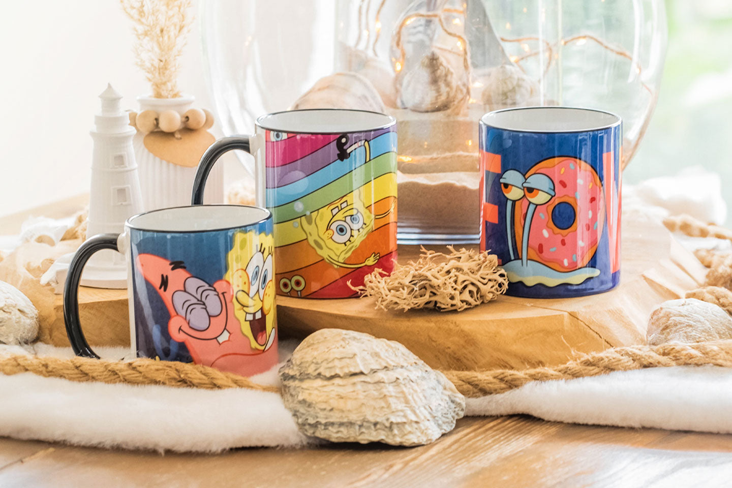 Drei SpongeBob Schwammkopf Tassen mit Motiven von SpongeBob, Patrick und Gary in einer hellen, maritim dekorierten Umgebung mit Muscheln und Leuchtturm.